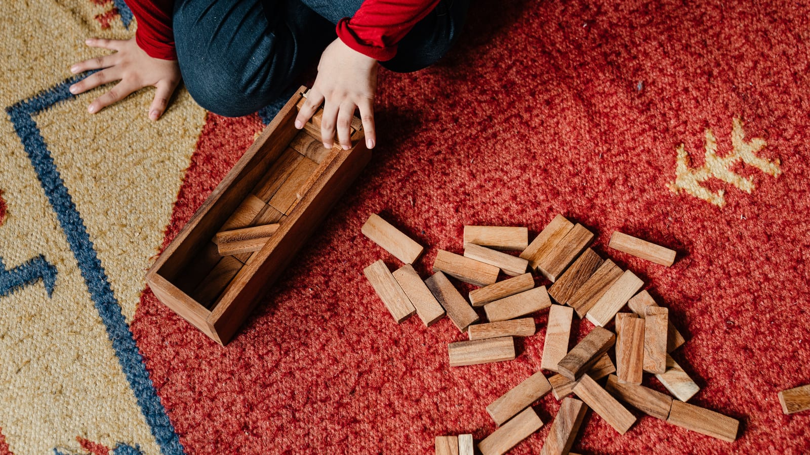 From above crop kid in casual clothes sitting on floor carpet while playing in jenga tower game with wooden blocks improving mental skills and accuracy