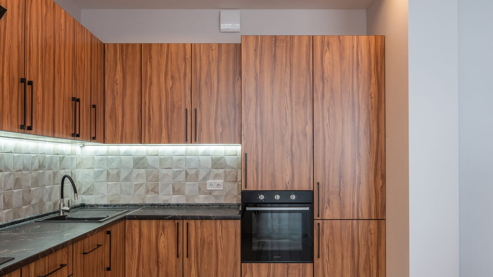 Interior of kitchen with wooden cupboards and contemporary black oven near marble counter with sink and tiled wall in light apartment