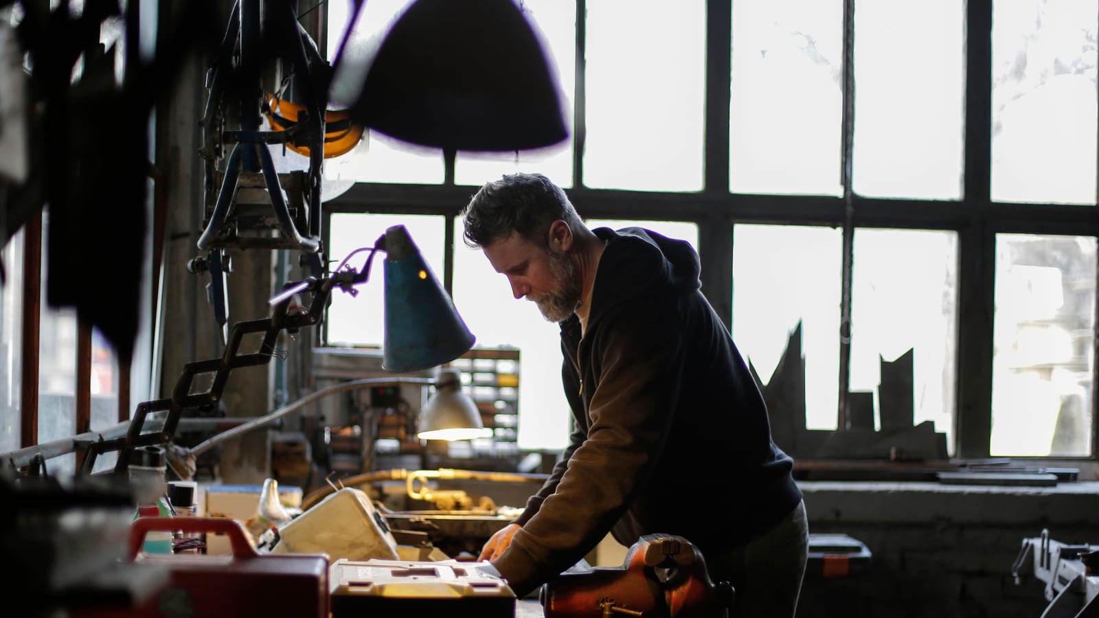 Side view of serious adult bearded male mechanic in workwear standing near workbench and working with details of engine in light of lamp in garage