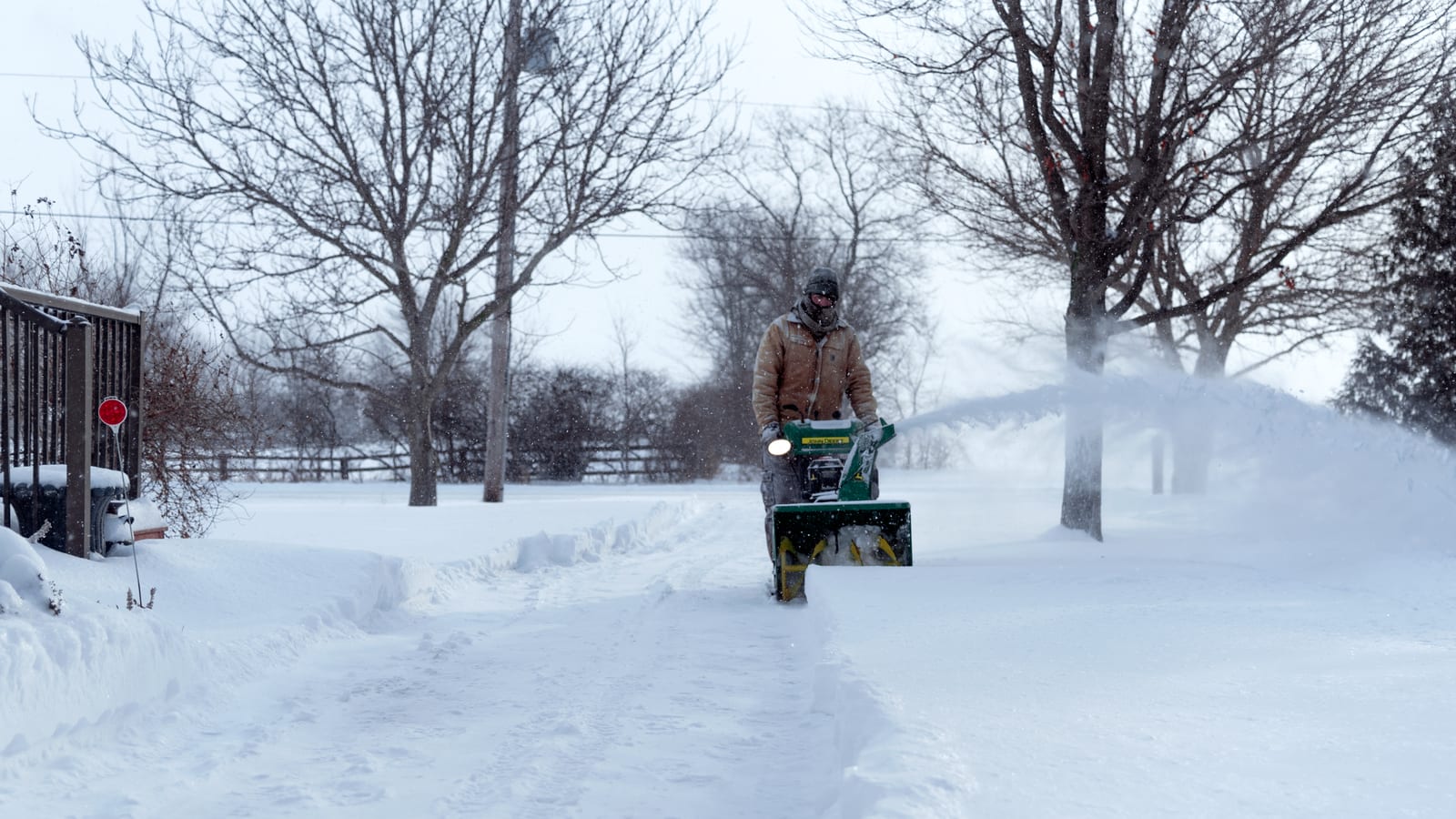 A person operates a snowblower on a snow-covered driveway in Essex, Ontario, during winter.