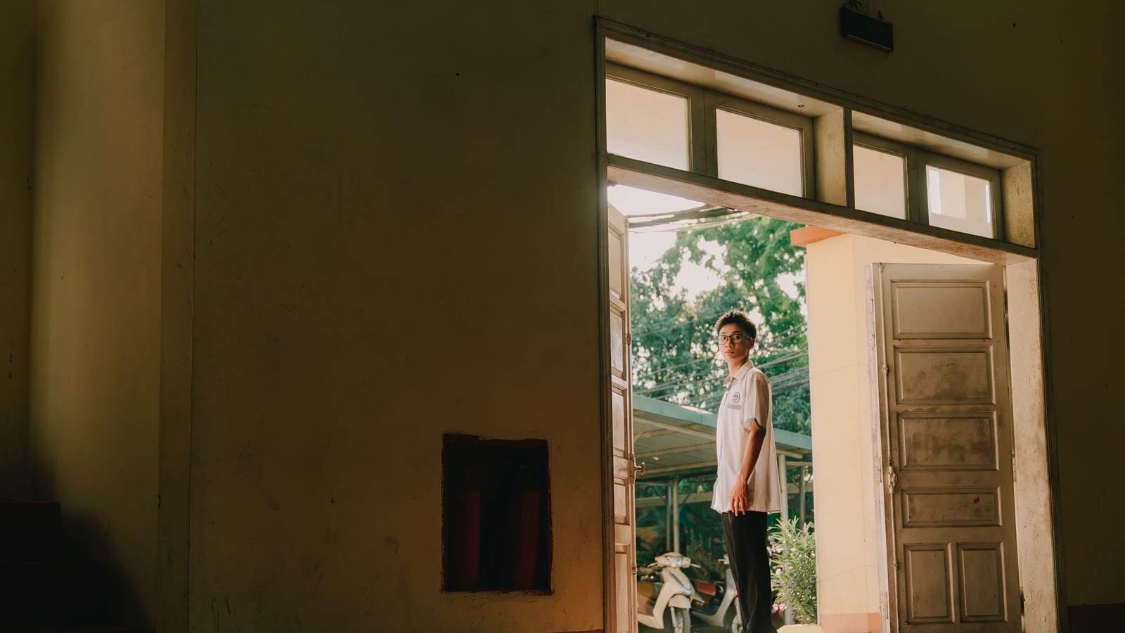 A young adult standing in a sunlit doorway, blending indoor and outdoor environments.